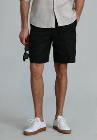 Shorts en lin noirs avec une coupe mi-longue, associés à des baskets blanches et une chemise à boutons de couleur claire. Tenant des lunettes de soleil dans une main.