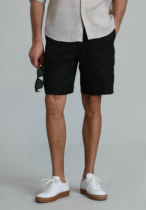 Shorts en lin noirs avec une coupe mi-longue, associés à des baskets blanches et une chemise à boutons de couleur claire. Tenant des lunettes de soleil dans une main.