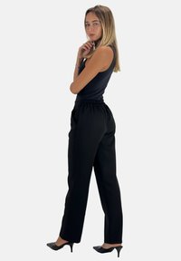 Zwarte elastische taillebroek met een rechte snit, gecombineerd met een aansluitend zwart tanktop. Het model draagt zwarte puntige hakken.