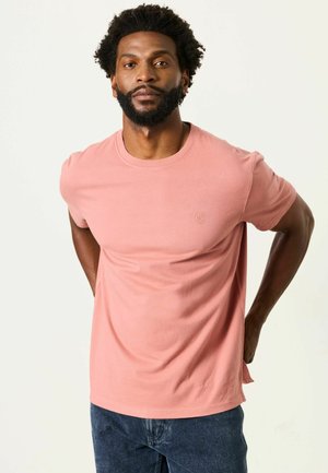 Mann mit kurzen lockigen Haaren und Bart, der ein einfarbiges pinkes T-Shirt und dunkelblaue Jeans trägt, stehend mit den Händen hinter dem Rücken.