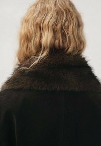 Cappotto scuro e morbido con un ampio colletto di pelliccia, caratterizzato da una texture liscia e un design minimalista. Lo sfondo è neutro e poco invadente.