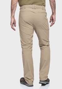 Beige technische Hosen haben einen geraden Schnitt, leicht zugängliche Gesäßtaschen und ein kleines Logo nahe dem Saum, kombiniert mit robustem Schuhwerk.