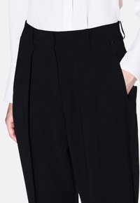 Zwarte high-waisted broeken met een op maat gemaakt ontwerp, plooien, zijzakken en een gladde, textuurstof. Gecombineerd met een wit shirt.