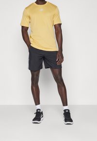 Żółta koszulka sportowa z krótkim rękawem i okrągłym dekoltem, z białym logo, zestawiona z czarnymi szortami z białymi paskami po bokach oraz czarnymi sneakersami.