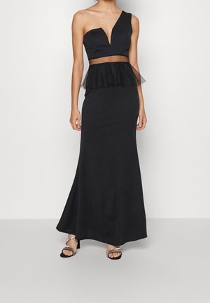 Vrouw draagt een lange zwarte jurk met één schouder, een doorschijnend taillepaneel en ruches, gecombineerd met zwarte sandalen met hoge hakken en strass-steentjes.