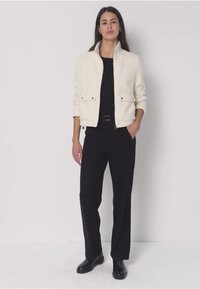 Nainen seisoo yksinkertaisen seinän vieressä valkoinen takki päällään mustan topin, mustien housujen ja mustien kenkien kanssa, toinen käsi taskussa.