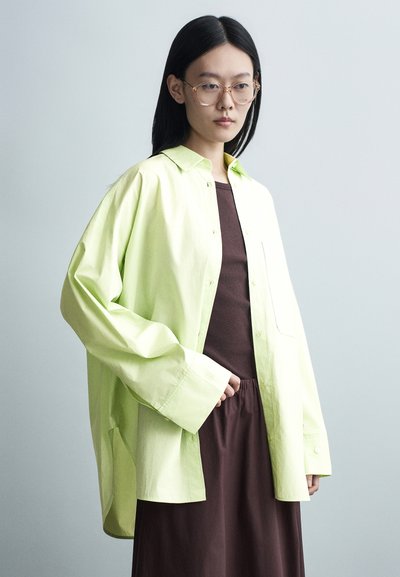 Jeune femme aux cheveux noirs raides, portant des lunettes à monture transparente, une chemise ample vert clair boutonnée sur une robe marron foncé, debout de profil.