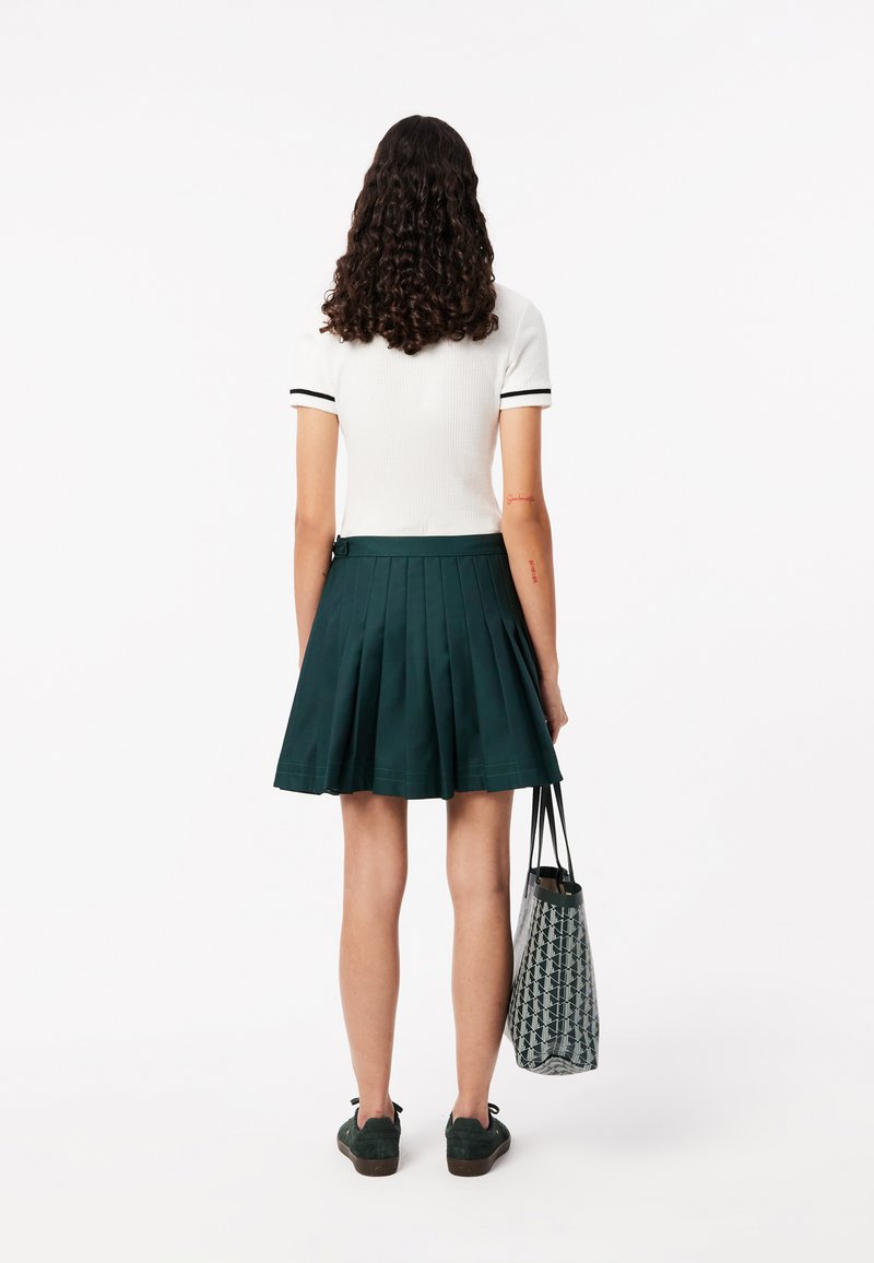 Weißes kurzärmliges Oberteil mit schwarzen Akzenten, kombiniert mit einem grünen plissierten Rock. Das Outfit beinhaltet schwarze Schnürschuhe und eine gemusterte Tragetasche.