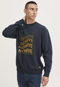 Sudadera azul marino con el texto en negrita y variado "ARCHIVE" en tonos dorados y verdes, cuello redondo y puños de canalé. Ajuste casual con detalle de etiqueta en el dobladillo.