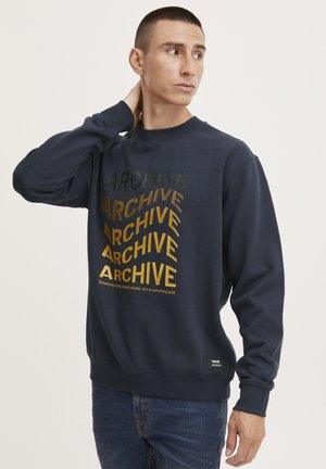 Navyblauwe sweatshirt met opvallende, variërende tekst "ARCHIVE" in gouden en groene tinten, ronde hals en geribde manchetten. Casual pasvorm met labeldetail aan de zoom.