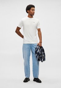 Balts īsām piedurknēm T-krekls ar nelielu teksta detaļu, brīvi pieguļoši zilie džinsi un zils kvadrātveida krekls, kuru tur vienā rokā. Melni apavi.