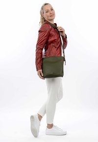 Olive green leather handbag, rectangular shape with textured surface and adjustable strap, positioned on shoulder, paired with a red jacket.