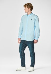 Camicia azzurro chiaro con maniche lunghe, abbinata a jeans blu scuro e sneaker bianche. Presenta un logo ricamato sul petto.