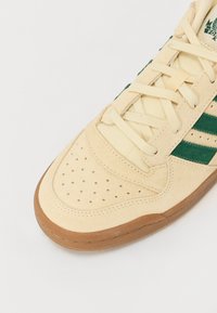 Beige sneakers med grønne striper, teksturert semsket skinn overdel, perforert tåområde og såle av gummigummi. Inkluderer flate lisser og polstret krage.