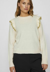 Kremowy sweter dziergany z ribbowanymi mankietami, z falbaniastymi akcentami na ramionach wykończonymi zielonym obszyciem. Luźny krój, miękka faktura i krótka długość.