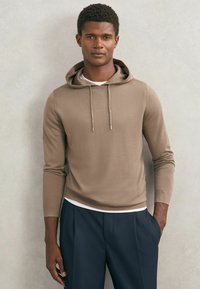 Ung mand med kort krøllet hår iført beige hættetrøje over hvid skjorte og marineblå bukser, stående mod en lysegrå væg.