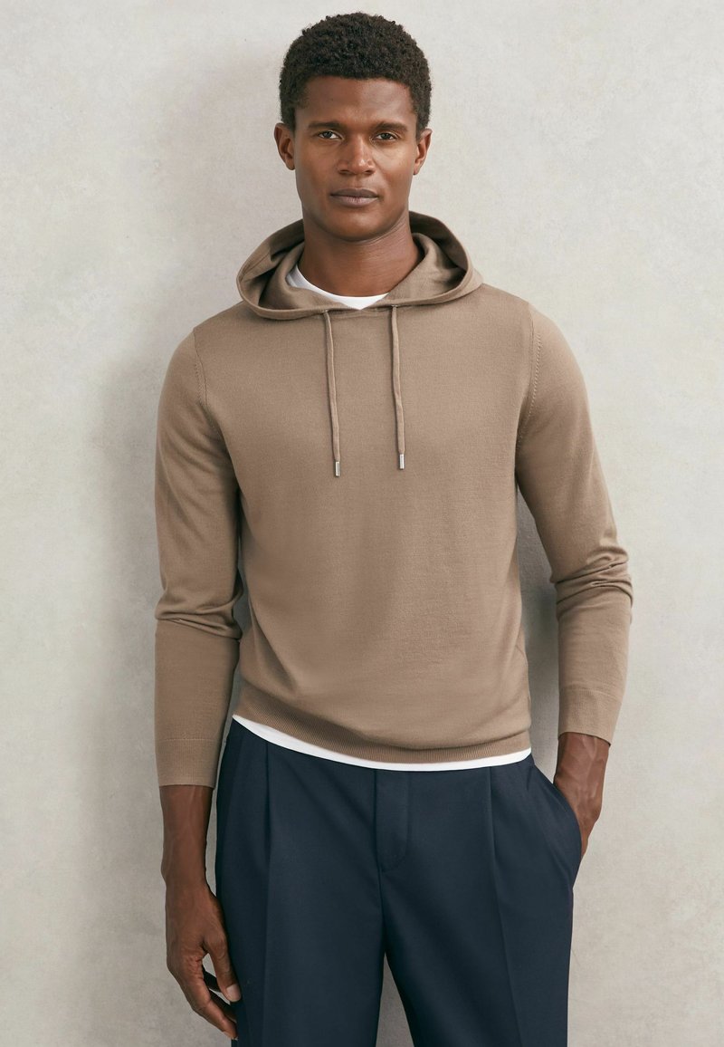 Ung mand med kort krøllet hår iført beige hættetrøje over hvid skjorte og marineblå bukser, stående mod en lysegrå væg.