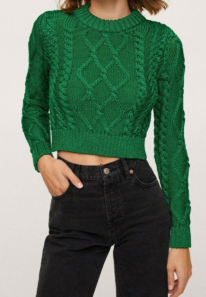 Femme en pull en maille vert texturé et court, et jean noir taille haute, une main dans la poche, sur un fond neutre.
