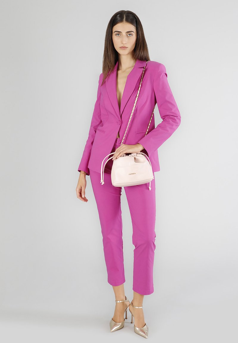 Blazer rosa e pantaloni abbinati con un design aderente. Tiene in mano una piccola borsa a sacchetto rosa chiaro. Sandali con tacco metallici.