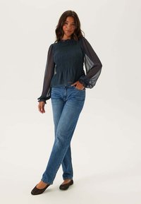 Námořnický modrý top s průhlednými dlouhými rukávy, nařaseným tělem a volánkovým výstřihem, spárovaný se světle modrými džíny a černými balerínami.