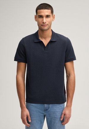 Uomo in piedi che indossa una polo a maniche corte di colore blu navy scuro con trama e jeans azzurri, su sfondo neutro.
