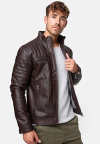Chaqueta de cuero marrón con detalles acolchados en los hombros, cuello alto y dos bolsillos con cremallera. Lleva puesta sobre una camiseta gris, diseño ajustado.
