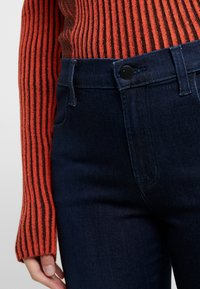 Vaqueros de mezclilla azul oscuro con un diseño ajustado, que presentan un cierre de botón negro y bolsillos frontales, combinados con un suéter a rayas rojas y negras.
