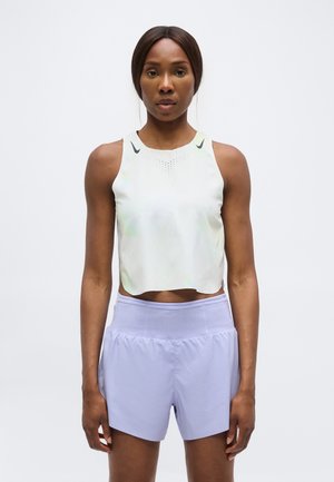 Femme portant un haut de sport blanc sans manches et un short de course violet clair, debout face à un fond uni.