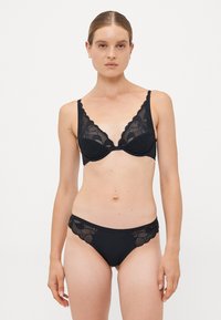 Vrouw met een lichte huid en opgestoken haar, gekleed in een bijpassende zwarte kanten bh en slipjes, staande tegen een effen lichte achtergrond.