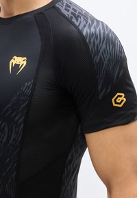 Κοντινό πλάνο ενός ατόμου που φοράει μαύρο αθλητικό πουκάμισο με πάνελ από δίχτυ, με χρυσό λογότυπο φιδιού στο στήθος και εξάγωνο λογότυπο στο μανίκι.