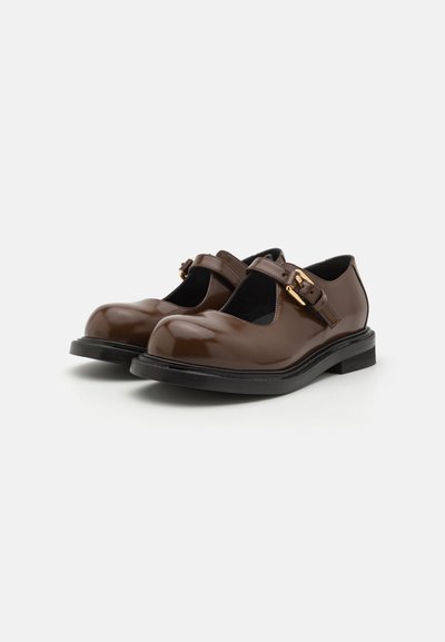 Zapatos Mary Jane de charol marrón con puntera redonda, correa ajustable y hebilla de tono dorado sobre una suela de goma negra.
