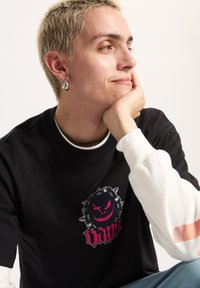 Camiseta negra con un gráfico colorido que presenta un círculo espinado y una cara siniestra. Se ven mangas largas blancas con un acento estampado.