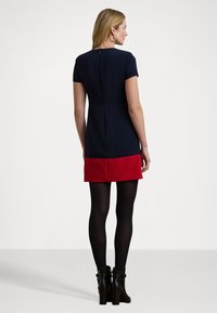 Vestido de mangas curtas azul-marinho com um painel inferior vermelho. Apresenta um fecho de correr nas costas e uma textura suave. Usado com collants pretos e botas de tornozelo.