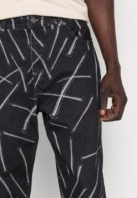 Black denim pants featuring a graphic pattern of white nails. The fabric has a textured appearance, with a standard fit and metal button closure.