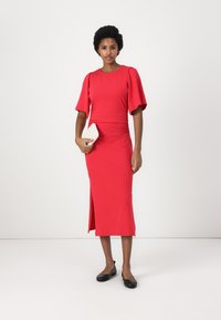 Abito midi rosso aderente con maniche corte e larghe e spacco laterale. Tessuto morbido e elastico. La modella tiene in mano una pochette crema e indossa ballerine nere.