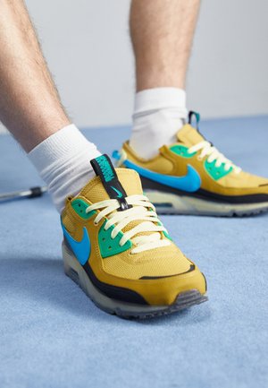 Persona che indossa sneakers Nike gialle e blu con lacci bianchi e calzini bianchi in piedi su un pavimento azzurro chiaro.
