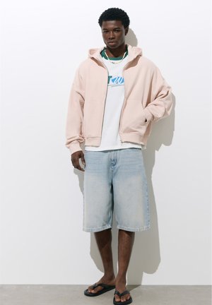 Jeune homme portant un sweat à capuche rose clair zippé, un t-shirt blanc à motif, un short en jean clair et des tongs noires, debout contre un mur blanc.