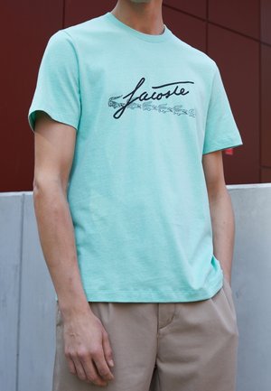 Mintgrünes Baumwoll-T-Shirt mit einem schwarzen, geschwungenen "Lacoste"-Logo und kleinen Krokodilgrafiken. Kurze Ärmel und Rundhalsausschnitt.