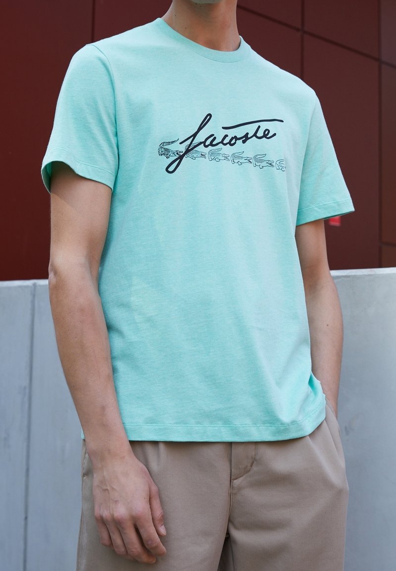 T-shirt en coton vert menthe avec un logo "Lacoste" en cursive noir et de petits graphiques de crocodile. Manches courtes et col rond.
