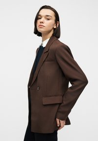 Νέος με κοντά σκούρα μαλλιά, που φοράει σκούρο καφέ σακάκι και πουκάμισο με γιακά, στέκεται και κοιτάζει με αυτοπεποίθηση προς το πλάι σε λευκό φόντο.