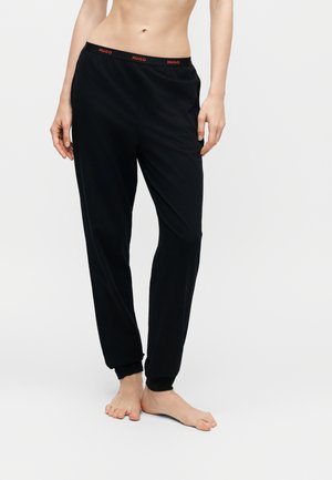 Personne portant un pantalon de jogging noir à taille élastique avec logo rouge "HUGO", debout pieds nus sur un fond clair uni.