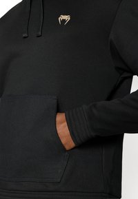 Μαύρο φούτερ με κουκούλα από υφή ύφασμα, με μπροστινή τσέπη, ριμπ μανσέτες και μικρό χρυσό λογότυπο κοντά στον λαιμό.