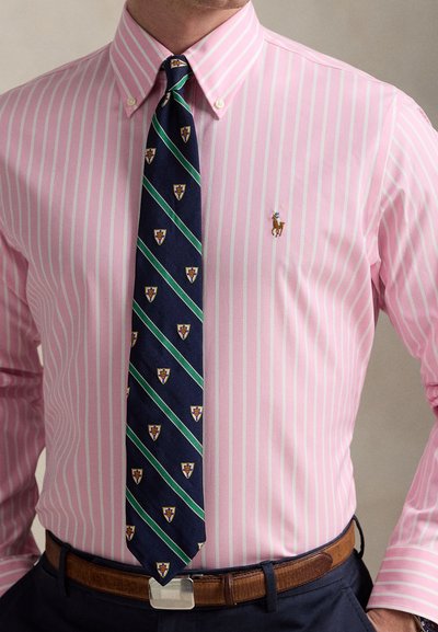 Ροζ ριγέ πουκάμισο με κολάρο που κουμπώνει, συνδυασμένο με μπλε γραβάτα με πράσινες διαγώνιες ρίγες και μοτίβα ασπίδων. Καφέ ζώνη ως διακοσμητικό στοιχείο.