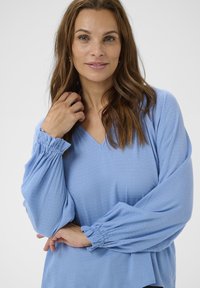 Bluză bleu deschis cu decolteu în V, mâneci bufante și manșete decolteu. Materialul are un model texturat discret pe toată suprafața. Croială largă și lejeră.