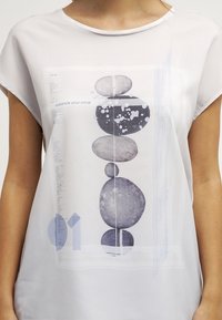 Blusa blanca de manga corta con un diseño impreso de piedras apiladas y elementos abstractos en tonos grises y azules. El texto dice "equilibra tu mente."