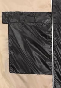 Schwarze Nylon-Tasche, die an einer beigen Stoffoberfläche befestigt ist, mit glatter Textur und sauberen Kanten sowie einem weißen Akzentbesatz entlang der Naht.