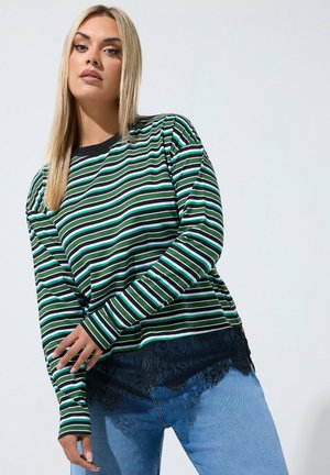 Vrouw met steil blond haar, gekleed in een groen, zwart en wit gestreept longsleeve-top met zwarte kanten afwerking en blauwe jeans tegen een effen achtergrond.