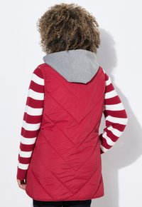 Chaleco aislante rojo con acolchado en forma de flecha, capucha gris y mangas a rayas blancas y rojas, con un corte largo y textura suave.