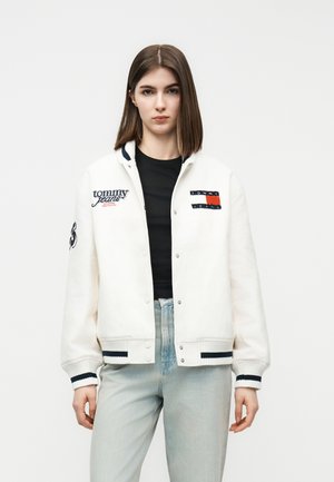 Junge Frau, die eine weiße Tommy Jeans Collegjacke über einem schwarzen Hemd und hellblauen Jeans trägt, vor einem schlichten Hintergrund stehend.
