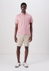 Πόλο μπλούζα σε ανοιχτό ροζ χρώμα, κοντομάνικη, με γιακά και κουμπάκια. Συνδυασμένη με μπεζ βερμούδα και γκρι-άσπρα αθλητικά παπούτσια.